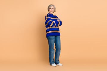 Confident mature woman wearing a blue striped sweater and jeans stands with arms crossed against a warm beige background