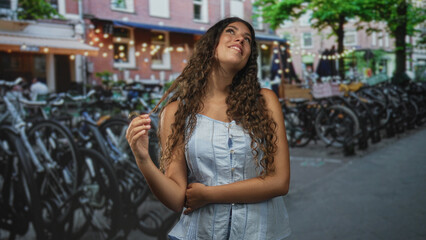 Hispanic teenage woman touching hair with right hand, smiling and looking up on a street with bicycle racks and cafe building; serenity confidence.