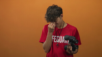 Teen lifeguard boy holding binoculars pinches nose and rubs eyes in studio with orange backdrop;...