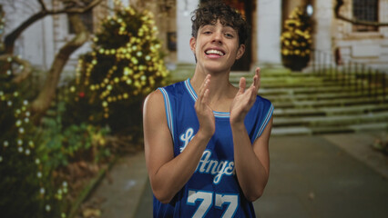 Man in blue los angeles basketball jersey clapping hands and smiling on steps of a building decorated with lights and trees; youthful joy.