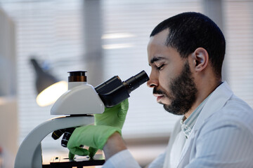 Young adult man examining specimen using laboratory microscope, wearing lab coat and green gloves,...