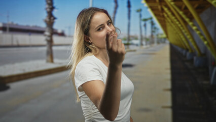 Young blonde woman beckons with hand toward camera on sunny urban street beside yellow canopy and palm trees; youthful confidence.