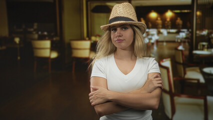 Woman wearing straw hat and white t shirt, arms crossed hugging her arm while standing in a dim...