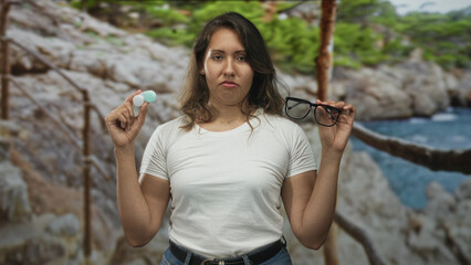 Woman holding glasses and contact lens case with eyes closed and a pained grimace, standing on a rocky seaside path near cliffs; disgust contact discomfort.