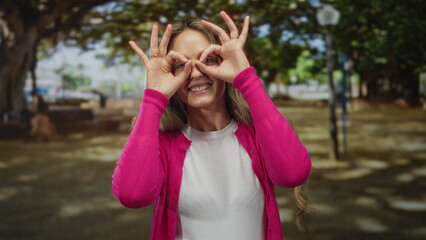 Naklejka premium Young blonde woman in pink cardigan making binoculars with hands in a sunny park outdoors, surrounded by lush green trees and cheerful atmosphere, enjoying nature playfully.
