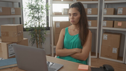 Young hispanic woman sitting at laptop by parcel stacks with arms crossed in delivery office; frustration.