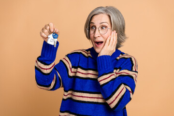 Aged mature beautiful woman with grey hair and glasses holds keys smiling in a blue striped sweater against a beige background suitable for real estate shopping lifestyle and wellness themes
