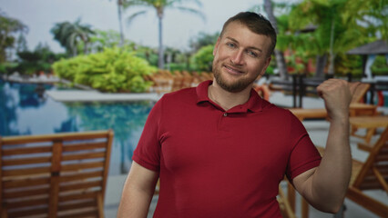 Fototapeta premium Man in red polo shirt points thumb toward resort pool deck while smiling; relaxed vacation leisure sunny.