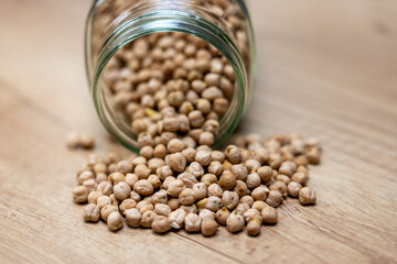 Dry chickpeas spilled from a glass jar on a wooden table. Natural vegan ingredient used in cooking, salads, soup and healthy meals.