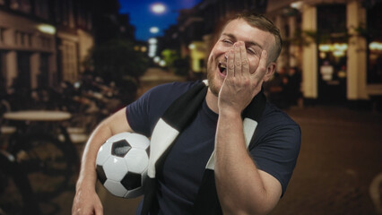 Man holds soccer ball and laughs, hand covering face, wearing black and white scarf in a city...