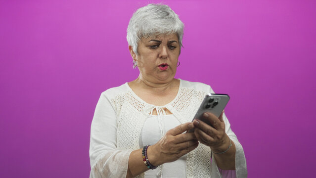 Senior woman grimacing while holding smartphone with hands and tapping screen in studio; confusion digital challenge. - Powered by Adobe