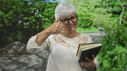Obraz premium Woman senior with grey hair holding open book with hands, smiling while reading and turning pages in forest path setting; learning contentment.