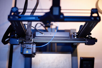 Closeup showing 3D printer operating with extruder head moving along rails, filament feeding through mechanism, advanced manufacturing technology in modern workspace