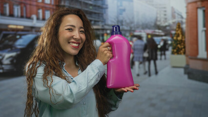 Smiling woman holding pink laundry detergent bottle on city street with storefronts and shoppers visible; household cleaning joy.