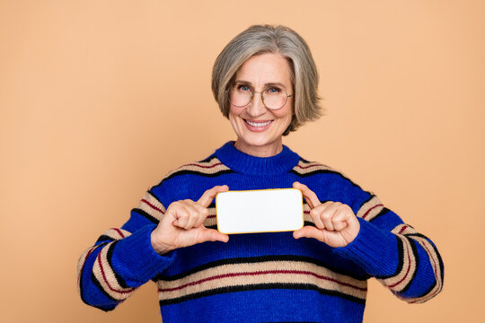 Aged mature woman with grey hair wear blue striped sweater smiles while hold blank smartphone screen beige background lifestyle fashion wellness shopping advertising