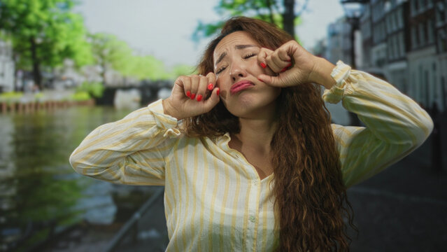 Young woman pulls her cheeks and pouts on a street in amsterdam beside a canal wearing a striped shirt; sulking playful humor.