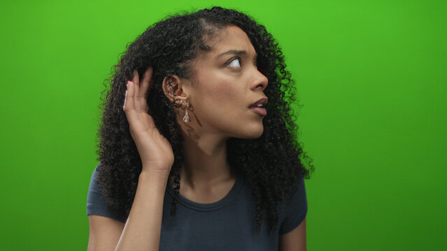 Woman holding hand cupped to ear wearing earrings listening on green studio set with subtle expression; curiosity. - Powered by Adobe