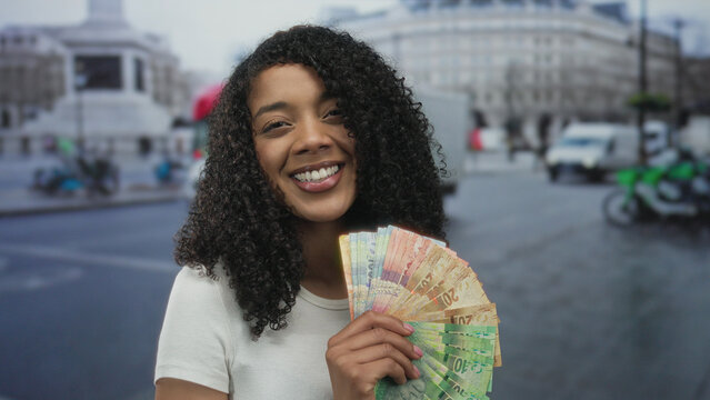 Woman smiling while holding rand banknotes on crowded city street in daylight; wealth ambition confidence.