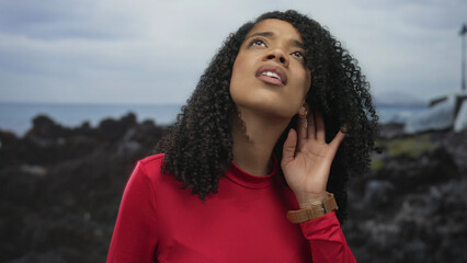 Woman holds hand to ear listening to ocean waves on rocky beach with head tilted upward and eyes closed under cloudy sky; curiosity.