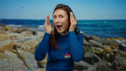 Young woman on a beach wearing a vote badge and blue sweater makes playful faces with hands near a scenic seaside backdrop.