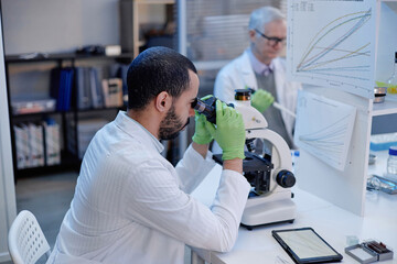Young adult man examining sample with microscope in laboratory while Caucasian man analyzing data on computer in background, scientific research environment, tablet on desk