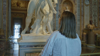 Brunette woman raises hand while looking at a marble sculpture in a museum building viewed from behind, wearing glasses; contemplation.