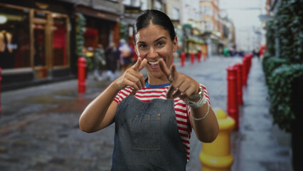Woman wearing denim apron and red striped shirt with hand to chest and pointing fingers up on a cobbled city street; gratitude joy.