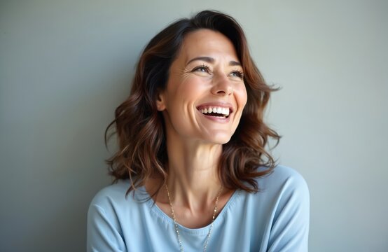 Happy middle-aged woman portrait smiling looking away. Attractive female with beautiful face expression, open mouth shows white teeth. Brunette lady wears blue shirt on grey studio background.