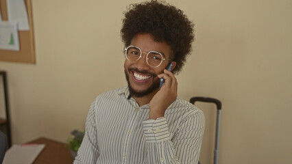 Smiling man talking on phone indoors in office wearing glasses with plant in background and striped shirt showing a cheerful and casual work environment.
