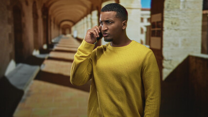Young black man holds smartphone to ear with tense, puzzled expression and furrowed brow in yellow sweater inside an arched building corridor, hands near face; concern decision.