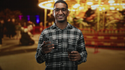Young black man smiling while holding a smartphone and coffee cup on a street near a carousel at night; joyful calm.