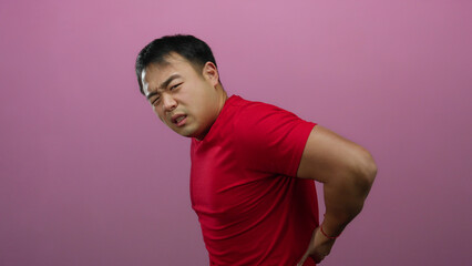 Young man in red shirt with back pain isolated against a vibrant pink wall, expressing discomfort as he grimaces, emphasizing health issues and body language.