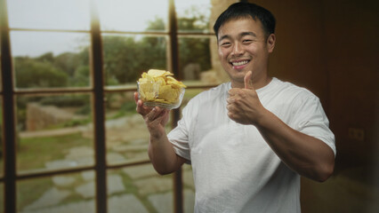 Man smiling points finger to transparent bowl filled with potato chips on wooden table in studio;...