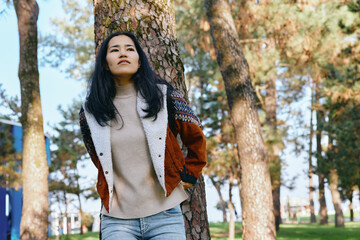 Fototapeta premium Woman outdoors in a park among trees, wearing a sweater and jacket, casual fashion and confident stance under sunlight, showcasing relaxed lifestyle in nature and scenery