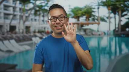 Man in blue t shirt smiling and waving hand at hotel resort pool area with sunbeds and palm trees;...