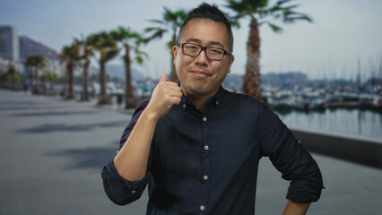 Man smiling gives thumbs up on seaside street by marina with palm trees and boats visible; optimism...