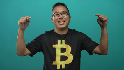 Man pointing fingers to bitcoin t shirt while smiling in a teal studio setting, hands visible and chest logo prominent; cryptocurrency pride.
