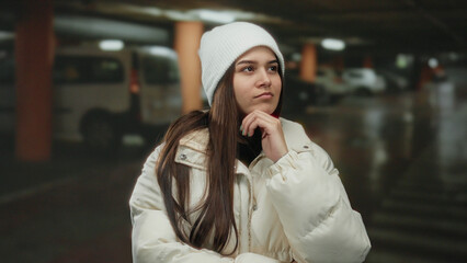 Fototapeta premium Young woman wearing a white jacket and beanie stands in an indoor parking lot with cars in the background, exuding a thoughtful expression.