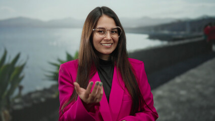Young woman gesturing on seaside promenade wearing pink blazer with ocean backdrop showing calm and relaxation
