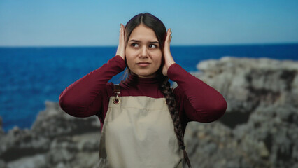 Hispanic waitress wearing an apron covers her ears by the seaside with a serene beach and sea background, capturing a moment of quiet introspection outdoors.