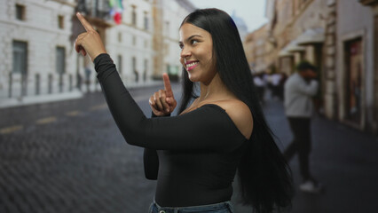 Fototapeta premium Smiling woman pointing finger upward on sunlit cobblestone city street showing bare shoulder; optimism confidence.