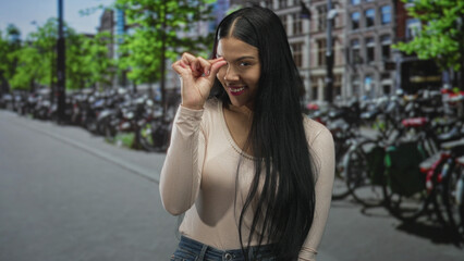 Young latin woman makes pinch gesture indicating small size on urban street bike parking area; playfulness.