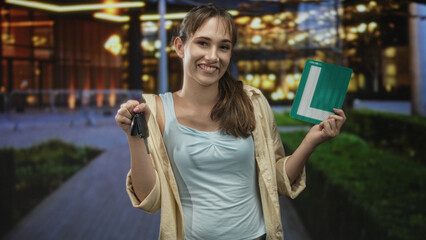 Young woman smiling holds car keys in one hand and a green learner plate in the other outside an illuminated building on a city street at night; pride new driver.
