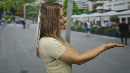 Smiling woman presenting on city street background, showcasing urban outdoor lifestyle with greenery and people in the distance, creating a lively urban scene.