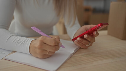 Woman writing notes while using smartphone in a new apartment, showcasing casual planning in her indoor living space.