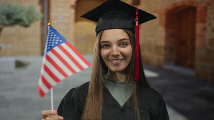 Young caucasian girl in black cap and gown holds small us flag beside weathered building facade;...