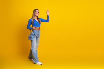 Naklejka premium Young woman in blue top and jeans waves hello against a bright yellow background for lifestyle fashion beauty and shopping appeal in advertising and promotional campaigns