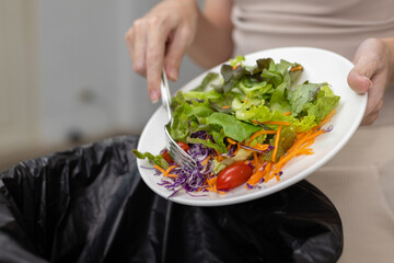 Close up of women discarding fresh vegetable salad into trash bin. Concept for food waste, diet rejection, meal dissatisfaction, eating disorder behavior or sustainability issue.