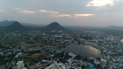 Aerial view of Pushkar Lake during the fair, capturing cloud reflections on the water, glowing roller-coaster lights, lakeside houses and ghats, with a warm sunset over the Aravali hills in the back