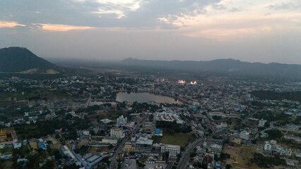 Aerial view of Pushkar Lake during the fair, capturing cloud reflections on the water, glowing roller-coaster lights, lakeside houses and ghats, with a warm sunset over the Aravali hills in the back